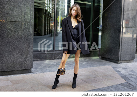 Young woman wearing elegant outfit oversize black blazer, Heeled Boots and mini dress, beautiful model stands, poses urban city. Modern high fashion, contemporary street style Young woman wearing elegant outfit oversize black blazer, Heeled Boots and mini dress, beautiful model stands, poses urban city. Modern high fashion, contemporary street style 121201267
