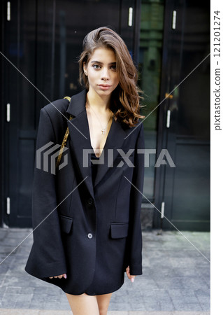Portrait young woman wearing elegant outfit oversize black blazer, beautiful model stands, poses confidently in urban city. Modern high fashion, contemporary street style on minimalist backdrop 121201274