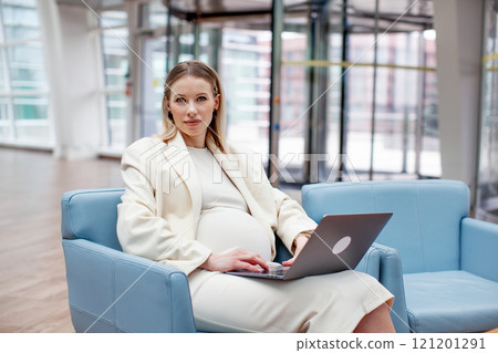 Beautiful pregnant businesswoman working indoors in office space with a laptop, dressed in fashionable white business attire consisting of a blazer and skirt. Modern motherhood into the corporate 121201291