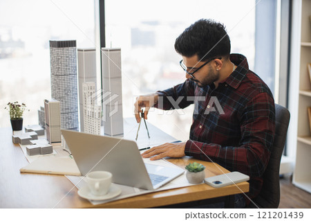Indian male architect in modern office measures city model showcasing creativity and precision in design process, highlighting architectural skills. 121201439