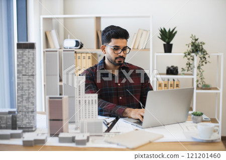 Young Indian male architect working on laptop, designing modern city plan with building models on desk. Young Indian male architect working on laptop, designing modern city plan with building models on desk. 121201460