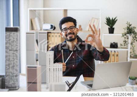 Young Indian male architect in office designing modern city architecture, gesturing okay symbol confidently at camera. Focus on creativity and innovation. Architectural models and laptop visible. Young Indian male architect in office designing modern city architecture, gesturing okay symbol confidently at camera. Focus on creativity and innovation. Architectural models and laptop visible. 121201463