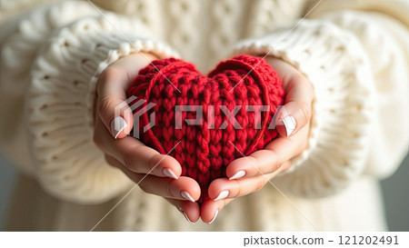 Female hands in a warm white sweater hold a red knitted heart 121202491