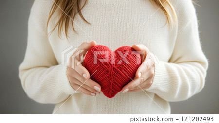 Female hands in a warm white sweater hold a red knitted heart 121202493