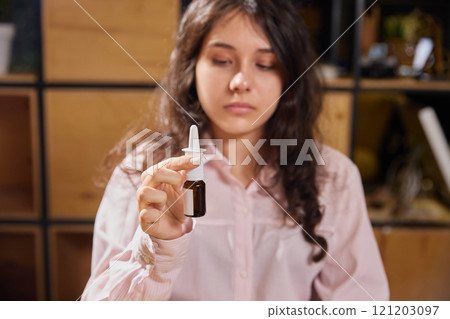 Young woman sitting at table, suffering from allergy, runny nose or nasal congestion in office and drops her nose with nasal spray 121203097