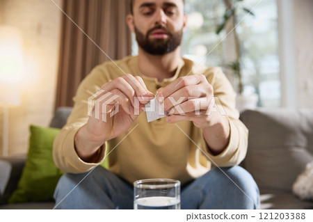 Close up of young sick man opens white sachet, preparing to take effervescent tablet while sitting at table on sofa in living room. 121203388