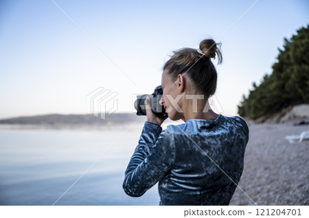 Over the shoulder view of a young woman photographer taking photos on a calm pebble beach 121204701