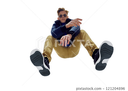 Low-angle bottom view image of fashionable young man wearing sunglasses, blue jacket, and mustard pants in dynamic pose. Urban style, modern, trendy look. Low-angle bottom view image of fashionable young man wearing sunglasses, blue jacket, and mustard pants in dynamic pose. Urban style, modern, trendy look. 121204896