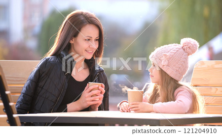 Young pretty woman having good time with her child daughter sitting at street cafe with hot drinks on sunny autumn day. Happiness in family relations concept 121204910