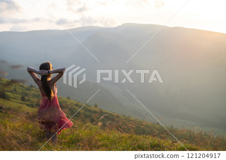 Young happy woman traveler in red dress standing on grassy hillside on a windy evening in summer mountains enjoying view of nature at sunset. 121204917
