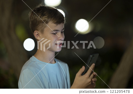 Teenage boy texting with his friend on cellphone outdoors on night city street 121205478