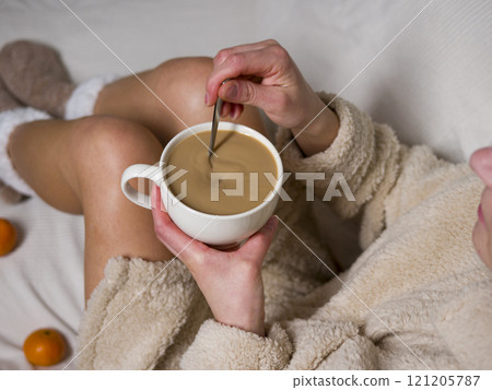Woman in cozy bathrobe sitting on bed, enjoying large cup of coffee while stirring it gently, with fresh tangerines nearby for peaceful morning Woman in cozy bathrobe sitting on bed, enjoying large cup of coffee while stirring it gently, with fresh tangerines nearby for peaceful morning 121205787