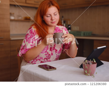 young woman working on tablet at home kitchen. A real female freelancer uses computer to work remotely from home with cup of morning coffee. 121206380