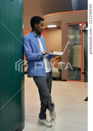 Vertical shot of laid back Black man dressed in striped blue shirt for office working on laptop keeping fingers on touchpad while standing against wall in coworking center, copy space 121206930