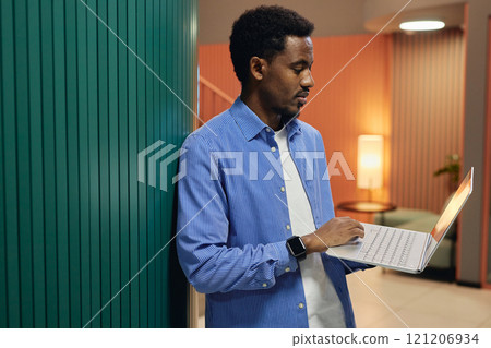 Side view of African American male employee dressed in striped blue shirt holding laptop typing on keyboard while standing against wall in coworking space, copy space 121206934