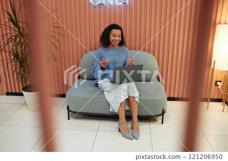 Full length shot of cheerful young multiethnic woman presenting during online conference joining video call via laptop, while sitting on comfy couch in lounge area of modern coworking space 121206950