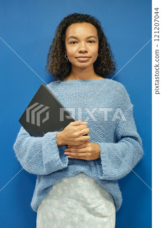 Vertical portrait of young multiethnic web designer dressed in elegant sweater holding laptop in hands looking confidently at camera while standing against blue wall Vertical portrait of young multiethnic web designer dressed in elegant sweater holding laptop in hands looking confidently at camera while standing against blue wall 121207044