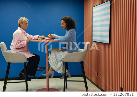 Side view of senior and younger female colleagues talking at cafe table developing design of new project while sitting on high chairs in modern coworking space, copy space Side view of senior and younger female colleagues talking at cafe table developing design of new project while sitting on high chairs in modern coworking space, copy space 121207051