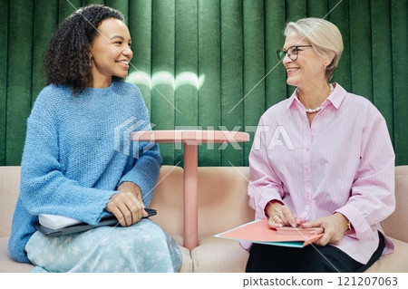 Smiling young multiethnic woman talking to senior female colleague exchanging work experience or assisting coworker with professional development, while sitting in lounge area of coworking space 121207063