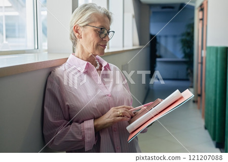 Medium shot of successful senior businesswoman wearing glasses reading note in notebook while standing in hallway of office building, copy space 121207088