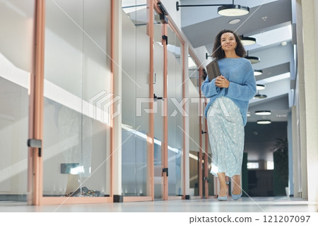 Full length shot of young multiethnic woman in elegant blue outfit holding laptop in hands walking along office rooms in hallway of modern coworking space, copy space 121207097
