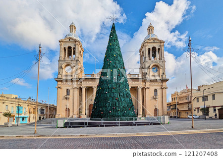 Christmas tree stands in front of historic Maltese church with twin bell towers, decorated for festive season under bright blue sky 121207408