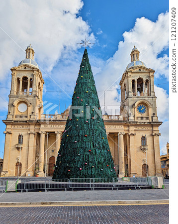Christmas tree stands in front of historic Maltese church with twin bell towers, decorated for festive season under bright blue sky 121207409