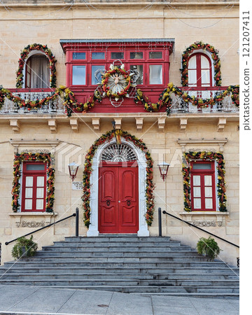 Decorated historic Maltese building with bold red door, adorned with colorful Christmas garlands and festive details Decorated historic Maltese building with bold red door, adorned with colorful Christmas garlands and festive details 121207411