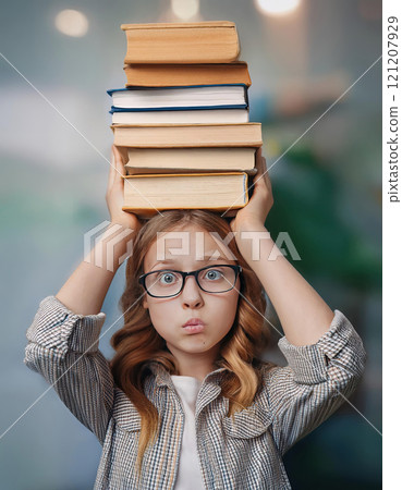 Fatigue pupil girl holds a pile of books on her head looks confused and bewildered due high volume of homework. Teenage student tired of difficult educational system 121207929