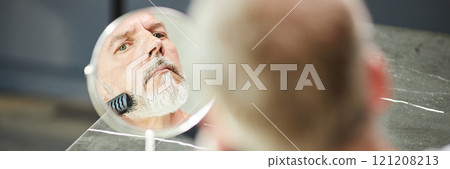 Website header shot of Reflection in small table mirror of mature man brushing neat beard during daily hair care routine at home, copy space 121208213