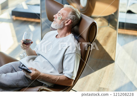 Medium shot of successful mature man moisturizing face skin with eye patches sitting in comfy armchair while relaxing with glass of red wine on weekend morning at home, copy space Medium shot of successful mature man moisturizing face skin with eye patches sitting in comfy armchair while relaxing with glass of red wine on weekend morning at home, copy space 121208228