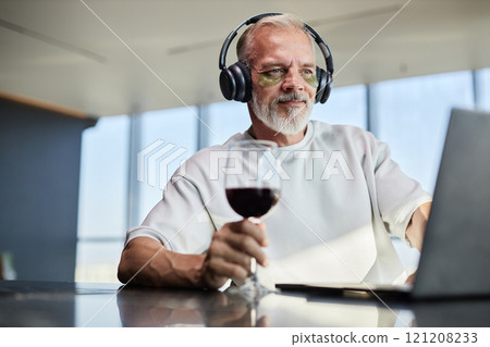 Medium shot of smiling mature man watching video on laptop wearing headphones while taking care of face skin with eye patches over glass of red wine at home, copy space 121208233
