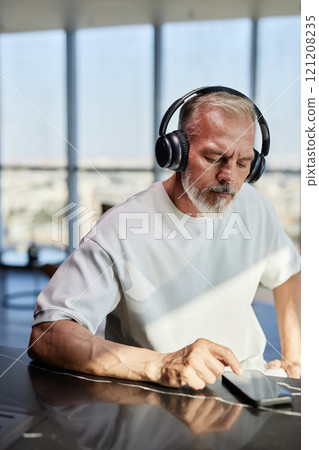 Vertical shot of mature man with neat beard text messaging on phone listening to music in headphones while sitting at table in contemporary apartment, copy space 121208235