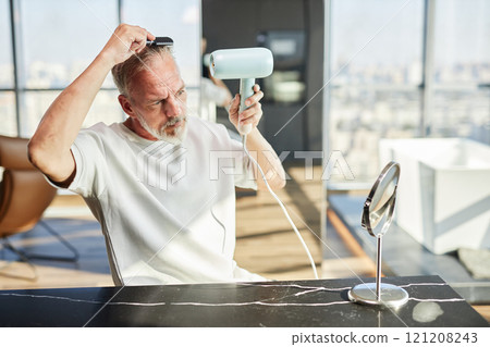 Medium shot of concentrated mature man sitting at table styling hair with hairdryer after taking shower during self care routine at home, copy space Medium shot of concentrated mature man sitting at table styling hair with hairdryer after taking shower during self care routine at home, copy space 121208243