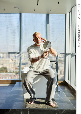 Full length shot of mature man with well groomed beard looking in hand mirror taking care of skin while sitting on bathtub in modern bathroom with panoramic windows, copy space 121208244