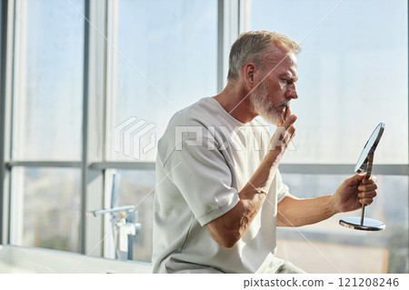 Side view of mature man with well groomed beard concerned about condition of facial hair looking in hand mirror while taking care of face in bathroom, copy space 121208246