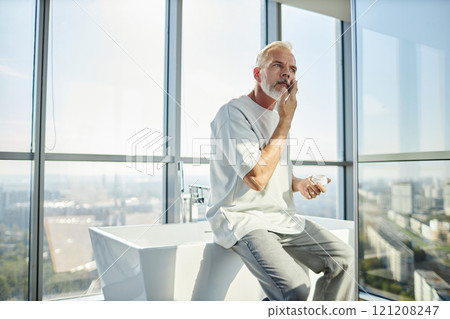 Medium shot of mature man in comfy home outfit applying nourishing cream on face taking care of skin while sitting on bathtub against big windows at home, copy space 121208247