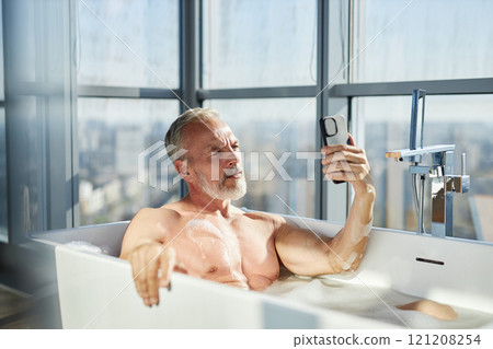 Medium shot of fit mature man with well groomed beard taking selfie lying in bathtub while relaxing in warm bath in bathroom with panoramic windows, copy space Medium shot of fit mature man with well groomed beard taking selfie lying in bathtub while relaxing in warm bath in bathroom with panoramic windows, copy space 121208254