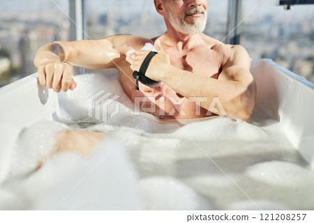 Cropped shot of fit mature man gently massaging skin with sponge taking relaxing foamy bath during body care routine in bathroom, copy space 121208257