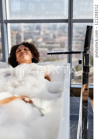 Vertical shot of smiling African American woman relaxing in bathtub filled with hot foamy water during self care ritual in room with panoramic windows 121208312