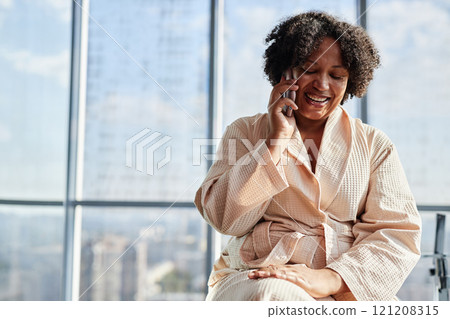 Medium shot of cheerful African American woman in comfy bathrobe talking on mobile phone during morning hygiene routine in bathroom with panoramic windows, slow life concept, copy space 121208315