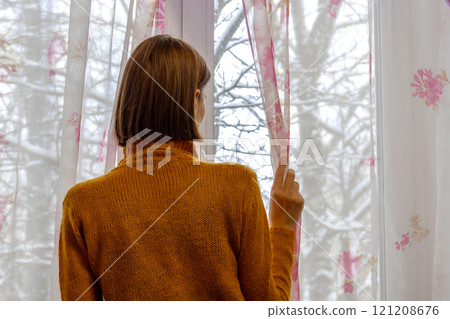 An unrecognizable woman in an orange sweater looks out window in winter. Rear view, close-up An unrecognizable woman in an orange sweater looks out window in winter. Rear view, close-up 121208676