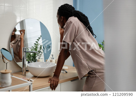 Back view of young African American woman standing at sink in bathroom looking in mirror while massaging face skin with jade roller at home, copy space Back view of young African American woman standing at sink in bathroom looking in mirror while massaging face skin with jade roller at home, copy space 121209590