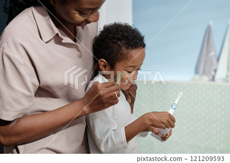 Candid shot of little Black boy laughing in mothers hands while woman massaging kids cheeks with jade roller in bathroom, happy family moment, copy space Candid shot of little Black boy laughing in mothers hands while woman massaging kids cheeks with jade roller in bathroom, happy family moment, copy space 121209593