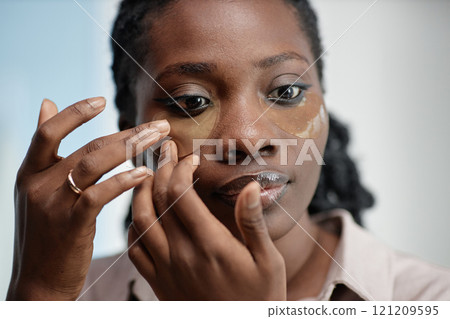 Portrait of young Black woman applying hydrogel patch under eye carefully lining mask with fingers during skin care rituals in bathroom Portrait of young Black woman applying hydrogel patch under eye carefully lining mask with fingers during skin care rituals in bathroom 121209595