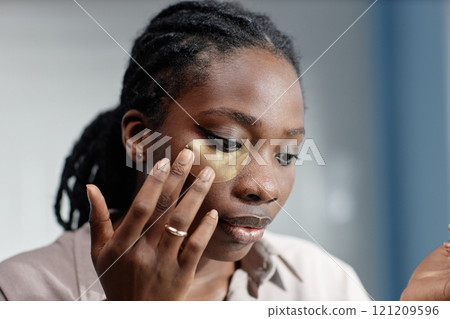 Portrait shot of young African American woman carefully applying hydrogel patch under eye lining mask with fingers while taking care of skin at home, copy space Portrait shot of young African American woman carefully applying hydrogel patch under eye lining mask with fingers while taking care of skin at home, copy space 121209596