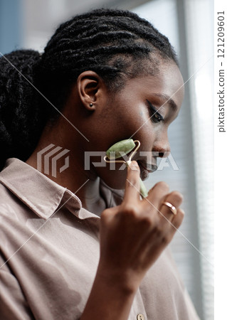 Profile of young African American woman massaging face with jade roller relaxing and cooling skin while looking in mirror in bathroom, copy space 121209601