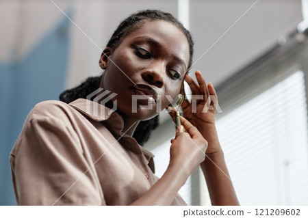 Relaxed young African American woman massaging face with jade roller soothing pores and cooling skin while looking in mirror at home, copy space 121209602