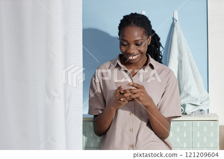 Medium shot of happy Black woman wearing cozy beige home suit text messaging on smartphone during self care routine in bathroom on Sunday morning, copy space Medium shot of happy Black woman wearing cozy beige home suit text messaging on smartphone during self care routine in bathroom on Sunday morning, copy space 121209604