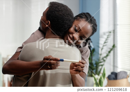 Medium shot of smiling young African American woman in husbands warm embrace holding pregnancy test while looking at positive result in bathroom 121209618